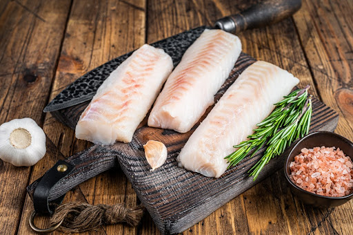 three raw cod loins sit on a cutting board on a wooden table.