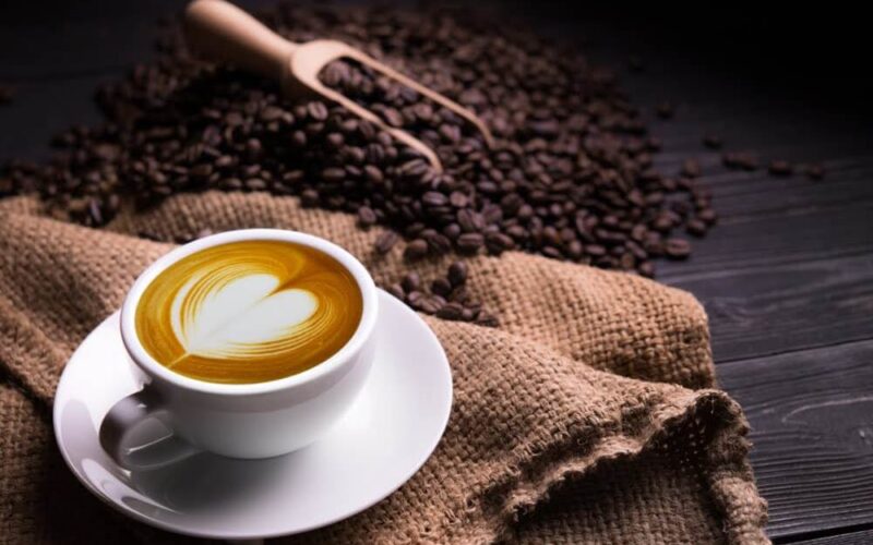 A latte in a mug sits on the edge of a table surrounded by a pile of coffee beans.